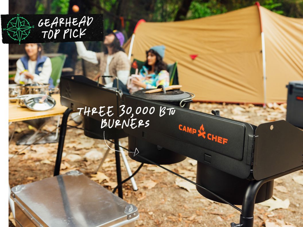 People sit in a campsite in front of a large tent. There is a three-burner stove in the foreground. Text overlay reads: Gearhead top pick, three 30,000 BTU burners.